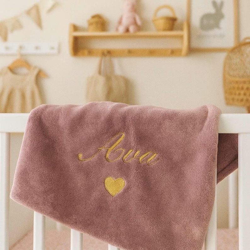 May include: A dusty rose baby blanket with the name "Ava" embroidered in gold script, along with a gold heart. The blanket is draped over a white crib rail. The nursery setting includes a wooden shelf with decor.