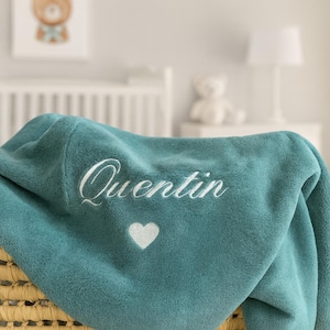 May include: Teal-colored baby blanket with the name "Quentin" embroidered in white script. A white heart is embroidered below the name. The blanket is folded over a woven basket, with a nursery scene in the background.