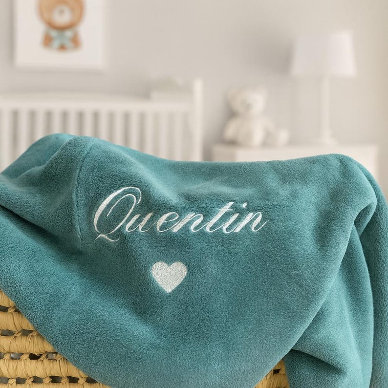 May include: Teal-colored baby blanket with the name "Quentin" embroidered in white script. A white heart is embroidered below the name. The blanket is folded over a woven basket, with a nursery scene in the background.