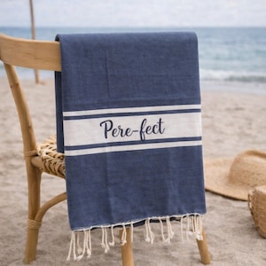 Peut inclure: Serviette de plage bleu marine avec des rayures blanches et le mot "Pere-fect" en écriture cursive. La serviette est drapée sur une chaise en bois sur une plage de sable. Un chapeau de paille et un panier tressé sont en arrière-plan.