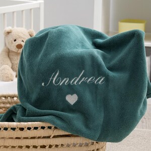 Può includere: Una coperta in pile verde acqua ricamata con il nome "Andrea" in corsivo bianco, insieme a un piccolo cuore bianco. La coperta è drappeggiata su un cestino di vimini, con un orsacchiotto visibile sullo sfondo.