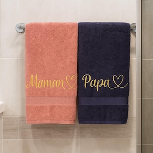 May include: Two bath towels hanging on a chrome bar. One is coral with "Maman" embroidered in gold script, the other is navy blue with "Papa" in gold script. Both have a heart symbol.