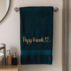 Peut inclure: Serviette de bain bleu canard avec l'inscription dorée "Papy bricolo" et un symbole de cœur brodé. La serviette est suspendue à un porte-serviettes argenté dans une salle de bain.
