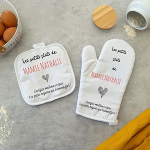 Peut inclure: Un ensemble de manique et de gant de cuisine blancs. Le texte "Les petits plats de MAMIE NATHALIE" est imprimé en noir et rouge. Un cœur noir est en dessous. Le texte "Certifiés meilleurs repas. Tes petits enfants qui t'aiment fort!" est imprimé en dessous.