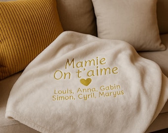 Puede incluir: Manta crema bordada con las palabras "Mamie On t'aime" en hilo dorado, un corazón dorado y nombres. La manta está sobre un sofá beige, con un cojín amarillo de pana y un cojín beige en el fondo. Un regalo afectuoso.