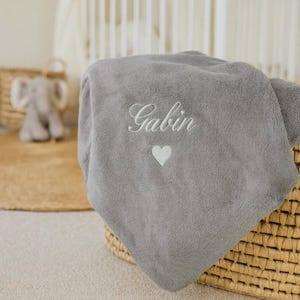 May include: A gray plush baby blanket embroidered with the name "Gabin" in cursive script and a small white heart. The blanket is folded and placed in a woven basket. A stuffed elephant toy is visible in the background.