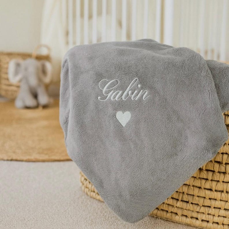 May include: A gray plush baby blanket embroidered with the name "Gabin" in cursive script and a small white heart. The blanket is folded and placed in a woven basket. A stuffed elephant toy is visible in the background.