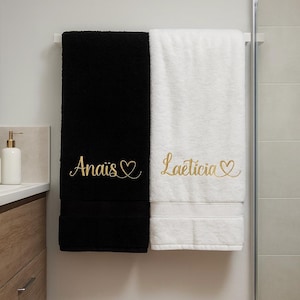 Peut inclure: Deux serviettes de bain rectangulaires suspendues côte à côte. L'une est noire unie et l'autre est blanche unie. Chaque serviette présente une broderie dorée en écriture cursive avec un cœur. La serviette noire porte le nom Anaïs et la blanche Laëticia.