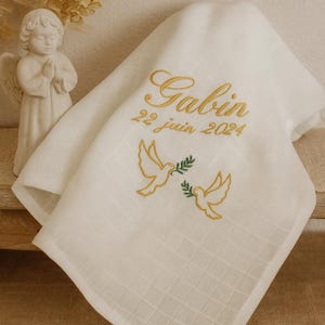 Peut inclure: Une couverture de bébé blanche, carrée, avec le texte brodé en or "Gabin" et "22 juin 2024". Deux colombes dorées avec des branches d'olivier sont brodées en dessous. La couverture est pliée.