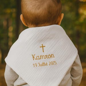 Peut inclure: Bavoir blanc avec une croix et le prénom "Kamron" brodés en fil doré, ainsi que la date "19 Juillet 2025". Le bavoir est porté par un bébé, dont on voit le dos de la tête. Fond flou.