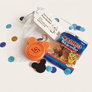 May include: A white sheer drawstring bag containing an orange circular keychain with a wave emblem, a black mouse-ear silhouette, and a small bag of Haribo Starmix sweets. A white tag attached reads: You've been Pixie-Dusted! From The Tomkins Family Stateroom 2706.