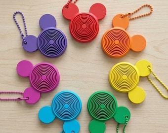 May include: A collection of seven circular keychains in red, orange, yellow, green, blue, purple, and magenta. Each keychain features a central concentric circle pattern and two smaller circular ears, with a matching coloured ball chain attached.