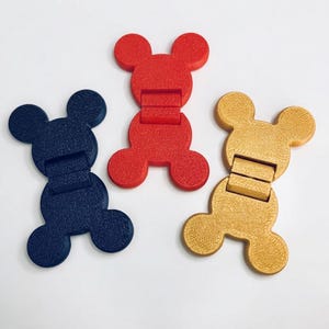 May include: Three hinged clips shaped like mouse heads in dark blue, red, and gold. Each clip features a textured surface and a central hinge mechanism that allows the two halves to fold together. The items are arranged in a triangular formation.