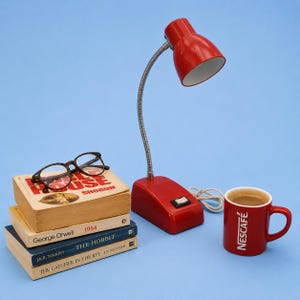 Puede incluir: Una lámpara de escritorio roja con cuello plateado flexible e interruptor en la base, junto a una pila de cuatro libros con gafas de montura oscura encima y una taza de cerámica roja con el logotipo blanco de Nescafé en el lateral.