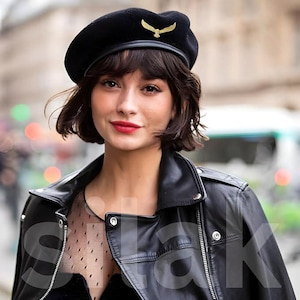 May include: A black beret with a gold-colored eagle emblem. The beret is worn with a black leather jacket. The person in the image has short, dark hair and red lipstick.