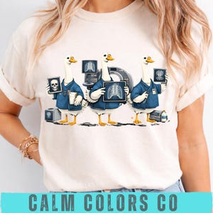Peut inclure: T-shirt crème avec trois oies de dessin animé en tenues médicales, chacune tenant une radiographie. Les oies sont devant un équipement médical. Le texte "CALM COLORS CO" est en bas du t-shirt.