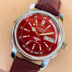 Puede incluir: Un reloj de pulsera automático Citizen con esfera roja y caja plateada. El reloj tiene una correa de cuero burdeos. La esfera muestra la fecha, la marca y el texto "21 Jewels" y "Japan Made".