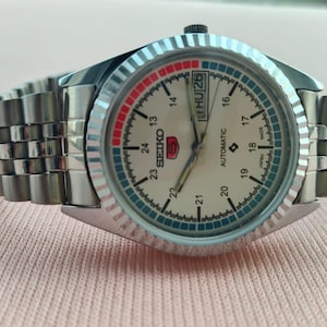 May include: A silver-toned Seiko 5 automatic watch with a cream-colored face. The watch features a day and date display, a rotating bezel with red and blue accents, and a stainless steel band. The watch is made in Japan.