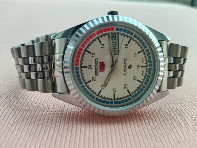 May include: A silver-toned Seiko 5 automatic watch with a cream-colored face. The watch features a day and date display, a rotating bezel with red and blue accents, and a stainless steel band. The watch is made in Japan.