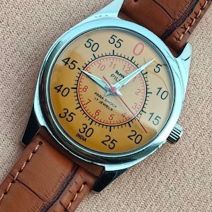 May include: A vintage wristwatch with a silver case and a tan face. The watch features black numerals, red accents, and a brown leather strap. The watch face reads "PARA SHOCK 17 JEWELS" and "INDIA".