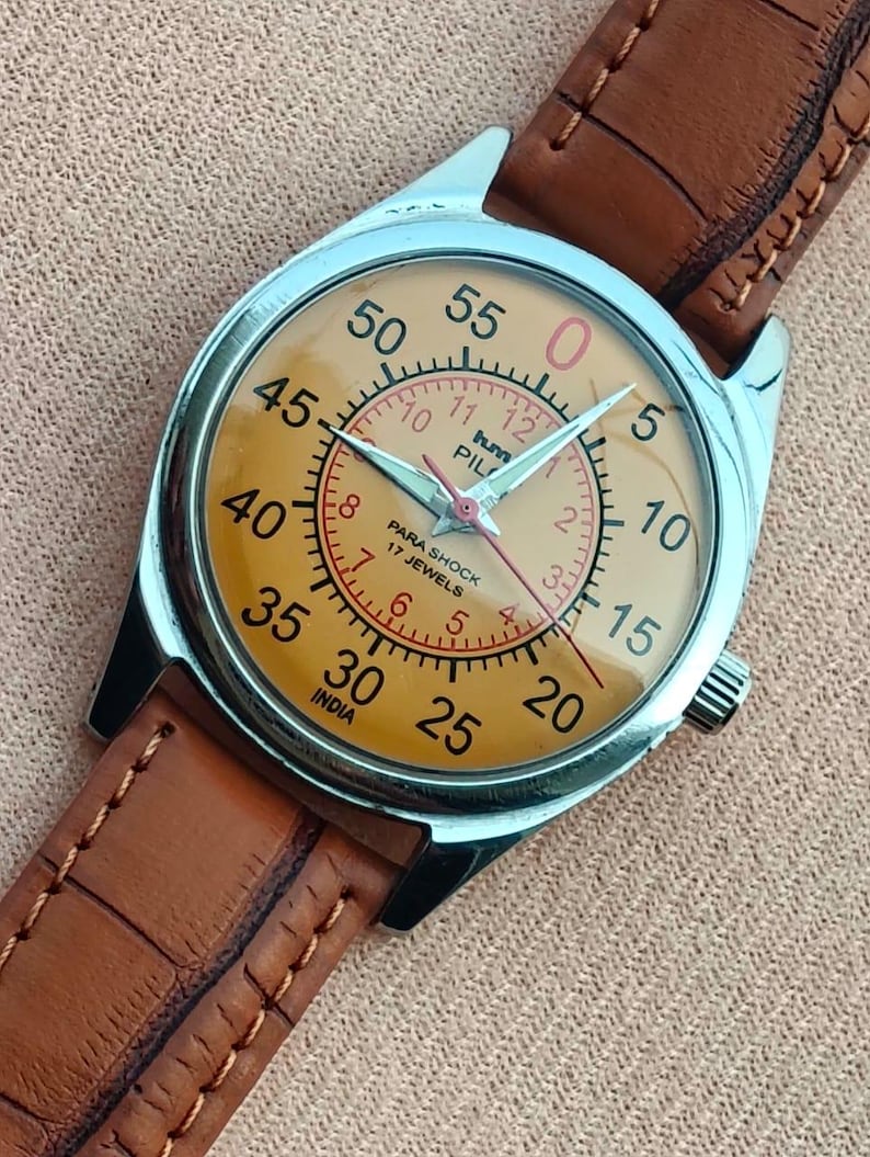 May include: A vintage wristwatch with a silver case and a tan face. The watch features black numerals, red accents, and a brown leather strap. The watch face reads "PARA SHOCK 17 JEWELS" and "INDIA".