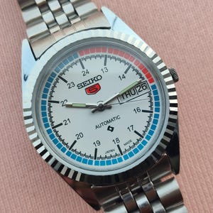 May include: A silver-toned Seiko 5 automatic wristwatch with a white face. The watch features a day and date display, a blue and red ring around the dial, and a silver link band. The watch face displays the text "SEIKO 5 AUTOMATIC JAPAN MADE".