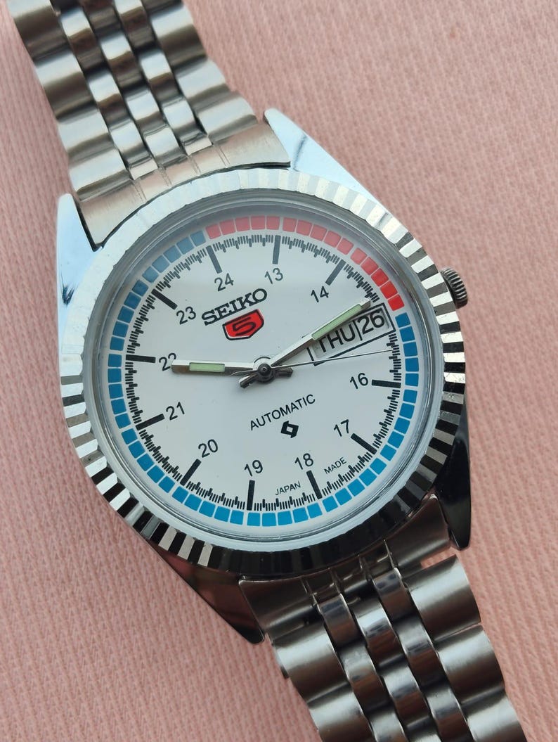 May include: A silver-toned Seiko 5 automatic wristwatch with a white face. The watch features a day and date display, a blue and red ring around the dial, and a silver link band. The watch face displays the text "SEIKO 5 AUTOMATIC JAPAN MADE".