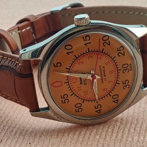 May include: A vintage wristwatch with a silver case and a tan leather strap. The watch face is orange with black numerals and red hands. The watch has the text "PARA SHOCK 17 JEWELS INDIA" on the face.