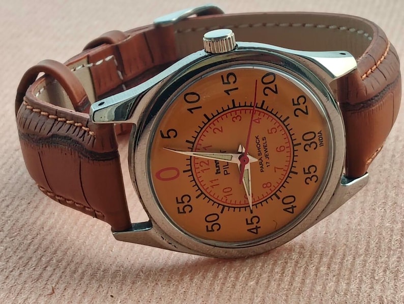 May include: A vintage wristwatch with a silver case and a tan leather strap. The watch face is orange with black numerals and red hands. The watch has the text "PARA SHOCK 17 JEWELS INDIA" on the face.
