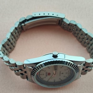 May include: A silver-toned Seiko 5 automatic wristwatch with a light-colored face. The watch features a fluted bezel and a stainless steel bracelet. The watch face displays the day and date.