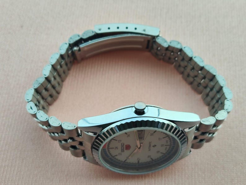 May include: A silver-toned Seiko 5 automatic wristwatch with a light-colored face. The watch features a fluted bezel and a stainless steel bracelet. The watch face displays the day and date.
