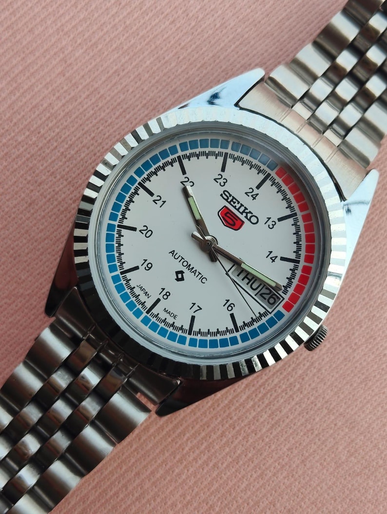 May include: A silver-toned Seiko automatic wristwatch with a white face, featuring a day-date display and a stainless steel band. The watch face has blue and red accents, with the text "SEIKO" and "AUTOMATIC". The watch is set to Thursday the 26th.