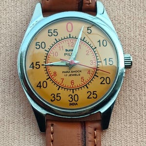 May include: A vintage wristwatch with a silver case and a tan face. The watch features a brown leather band and a dial with black and red numerals. The watch face includes the text "PARA SHOCK 17 JEWELS INDIA".