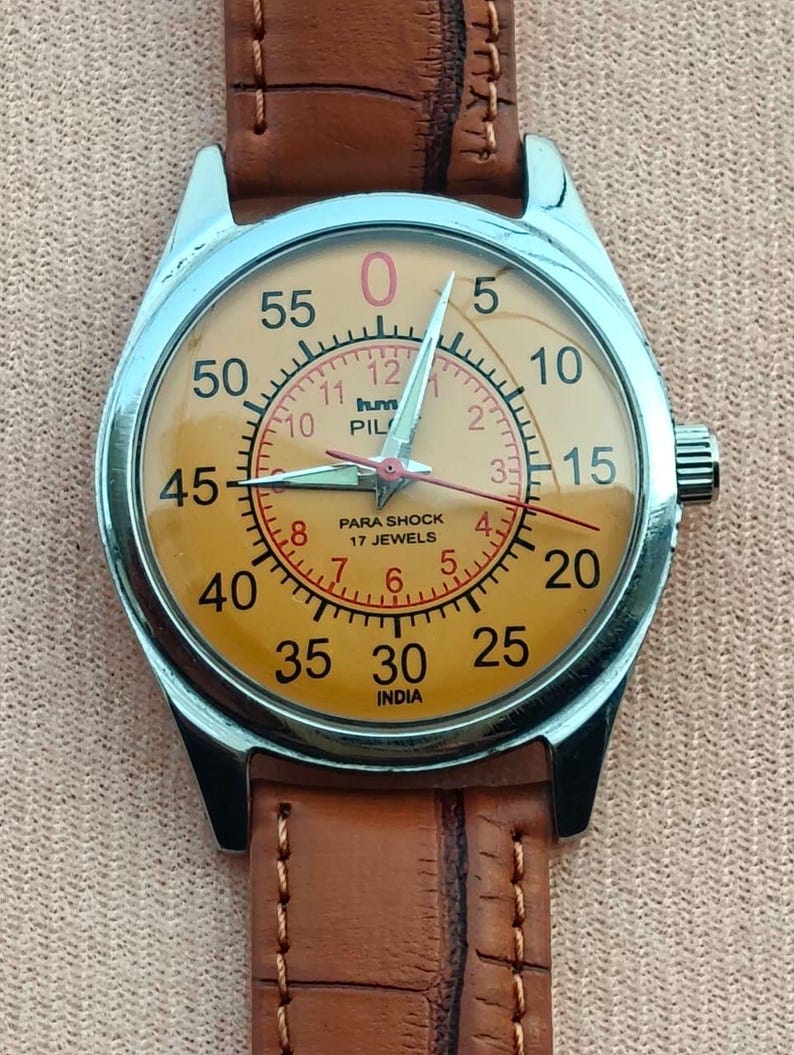 May include: A vintage wristwatch with a silver case and a tan face. The watch features a brown leather band and a dial with black and red numerals. The watch face includes the text "PARA SHOCK 17 JEWELS INDIA".
