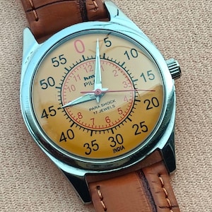 May include: A vintage wristwatch with a light orange face, silver case, and brown leather band. The watch face features black numerals, red accents, and the text "PILOT" and "INDIA". The watch has a classic design.