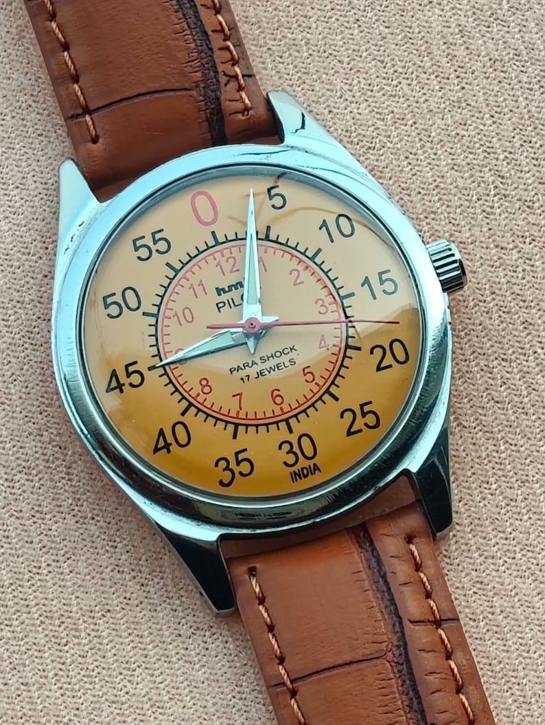 May include: A vintage wristwatch with a light orange face, silver case, and brown leather band. The watch face features black numerals, red accents, and the text "PILOT" and "INDIA". The watch has a classic design.