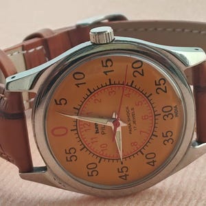 May include: A vintage wristwatch with a silver case and a tan leather band. The watch face is orange with black and red numerals and hands. The watch has the text "PARA SHOCK 17 JEWELS INDIA" on the face.