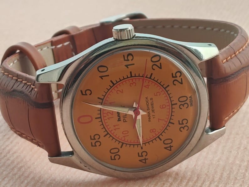 May include: A vintage wristwatch with a silver case and a tan leather band. The watch face is orange with black and red numerals and hands. The watch has the text "PARA SHOCK 17 JEWELS INDIA" on the face.