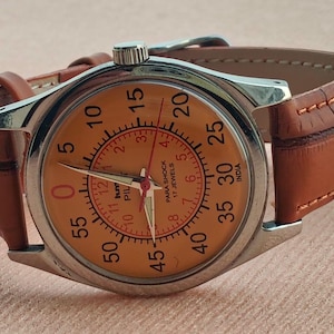 May include: A vintage wristwatch with a silver case and a tan face featuring black numerals and red accents. The watch has a brown leather band. The watch face reads "PARA SHOCK 17 JEWELS INDIA".