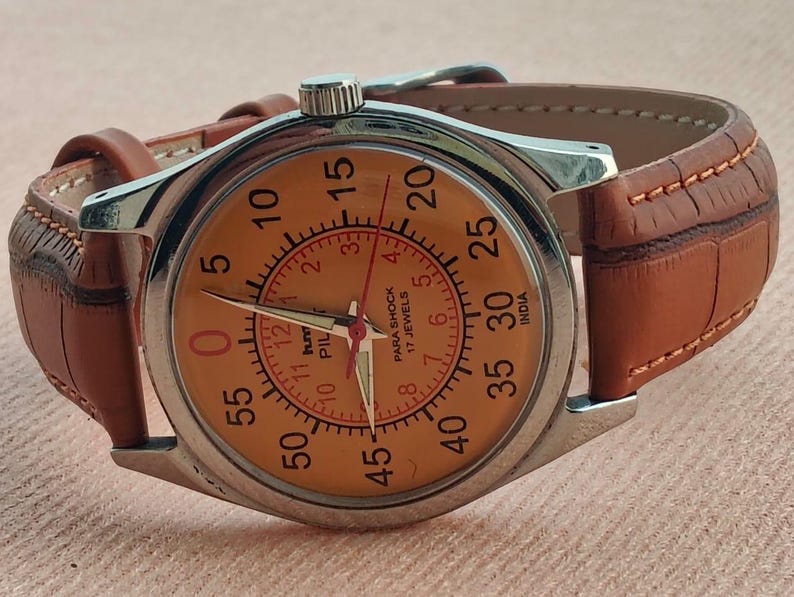 May include: A vintage wristwatch with a silver case and a tan face featuring black numerals and red accents. The watch has a brown leather band. The watch face reads "PARA SHOCK 17 JEWELS INDIA".