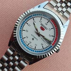 May include: A silver-toned Seiko automatic wristwatch with a white face, featuring a day-date display and a stainless steel band. The watch face has blue and red accents, with the word "SEIKO" and "AUTOMATIC" visible. The watch is set to Thursday the 26th.