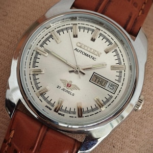 May include: A Citizen Automatic wristwatch with a silver dial, silver-tone hour markers, and a day-date window at the three o'clock position. The watch features a brown leather strap with a textured finish and a stainless steel case.