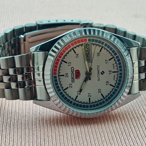 May include: A silver-toned Seiko 5 automatic wristwatch with a light-colored face, day and date display, and a stainless steel band. The watch face features red and blue accents and a fluted bezel.