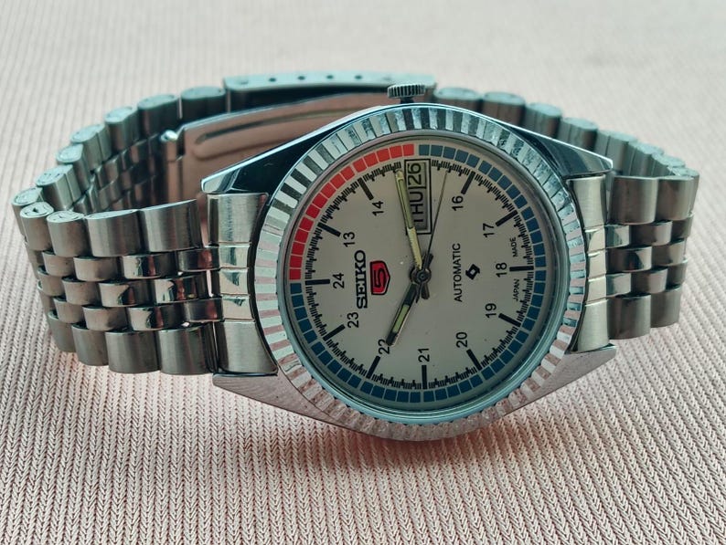 May include: A silver-toned Seiko 5 automatic wristwatch with a light-colored face, day and date display, and a stainless steel band. The watch face features red and blue accents and a fluted bezel.