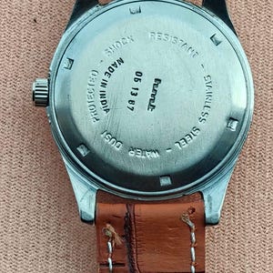 May include: Close-up of a vintage wristwatch with a brown leather strap. The watch face is stainless steel with the text "SHOCK RESISTANT" and "WATER DUST PROTECTED". The watch is marked "MADE IN INDIA" and has the date "05 13 87".