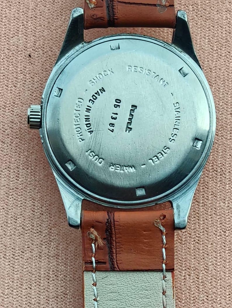 May include: Close-up of a vintage wristwatch with a brown leather strap. The watch face is stainless steel with the text "SHOCK RESISTANT" and "WATER DUST PROTECTED". The watch is marked "MADE IN INDIA" and has the date "05 13 87".