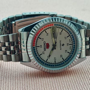 May include: A silver-toned Seiko 5 automatic wristwatch with a light-colored face, day and date display, and a stainless steel band. The watch face features red and blue accents and a fluted bezel.