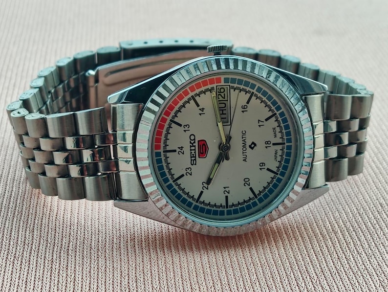 May include: A silver-toned Seiko 5 automatic wristwatch with a light-colored face, day and date display, and a stainless steel band. The watch face features red and blue accents and a fluted bezel.