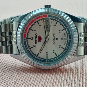 May include: A silver-toned Seiko 5 automatic wristwatch with a light-colored face. The watch features a day and date display, a fluted bezel, and a stainless steel band. The watch face displays the Seiko logo and the text "AUTOMATIC".