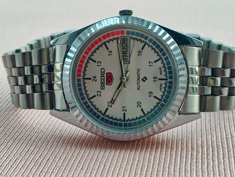 May include: A silver-toned Seiko 5 automatic wristwatch with a light-colored face. The watch features a day and date display, a fluted bezel, and a stainless steel band. The watch face displays the Seiko logo and the text "AUTOMATIC".